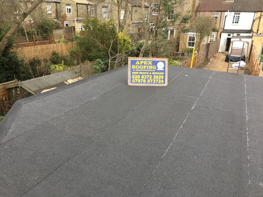 Flat roof with a sign for Apex Roofing and Maintenance in the center, advertising new roofs and repairs with contact numbers, surrounded by residential buildings and garden fences.
